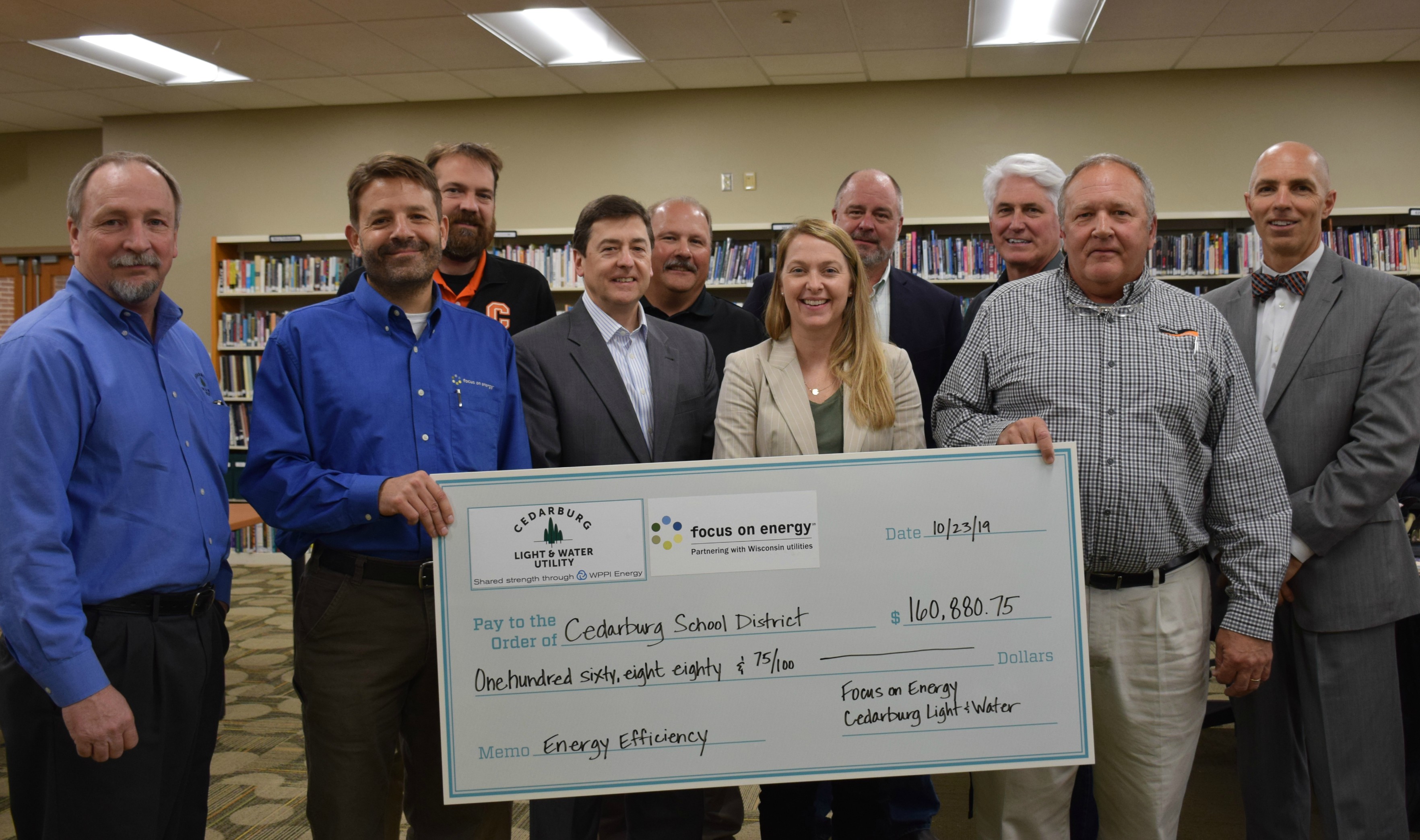 A group of professionally-dressed people stand in a room with bookshelves, holding a large check from Cedarburg Light & Water Utility and Focus on Energy.