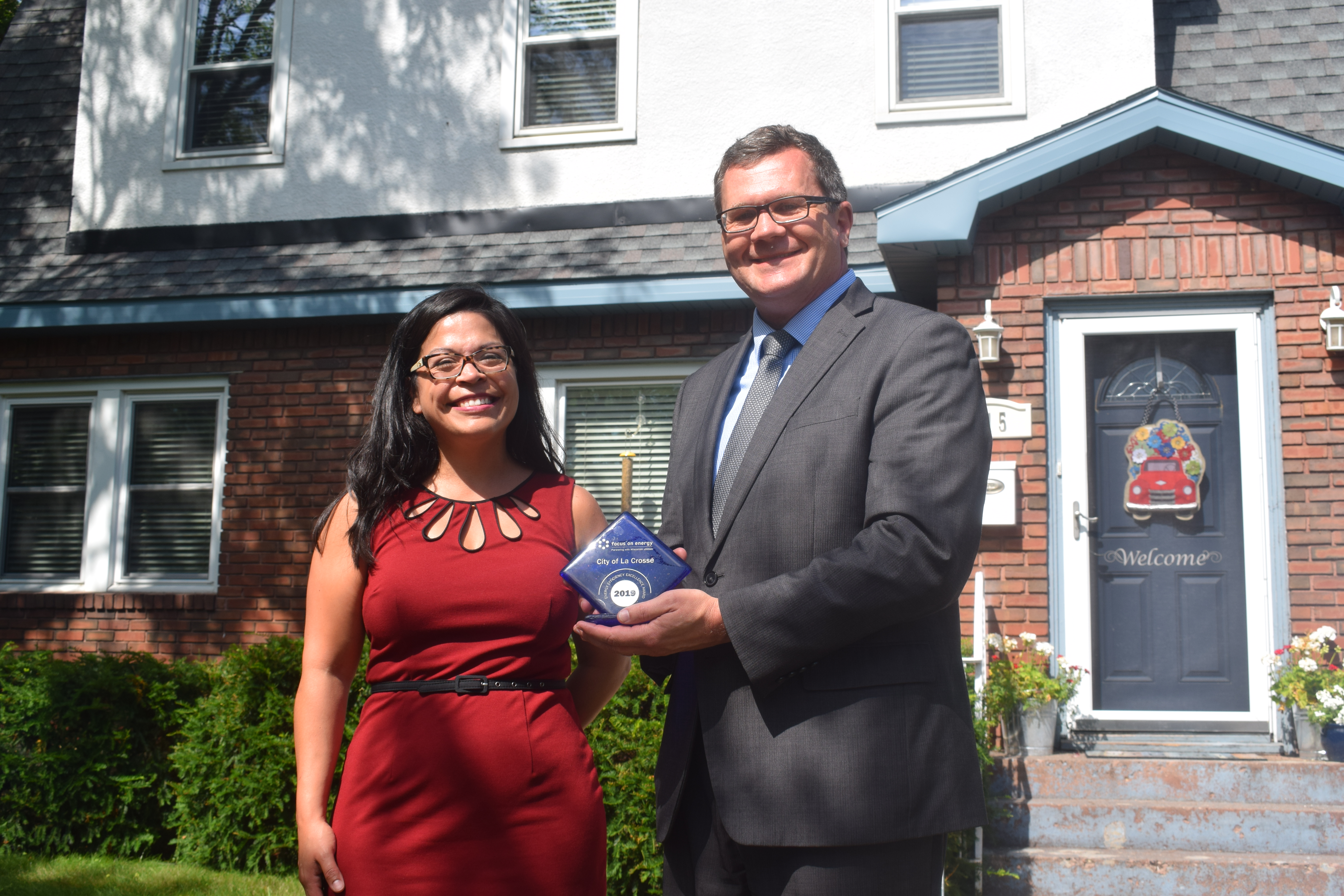 A man and a woman stand in front of a house holding the blue Focus on Energy award. It's a sunny summer day.