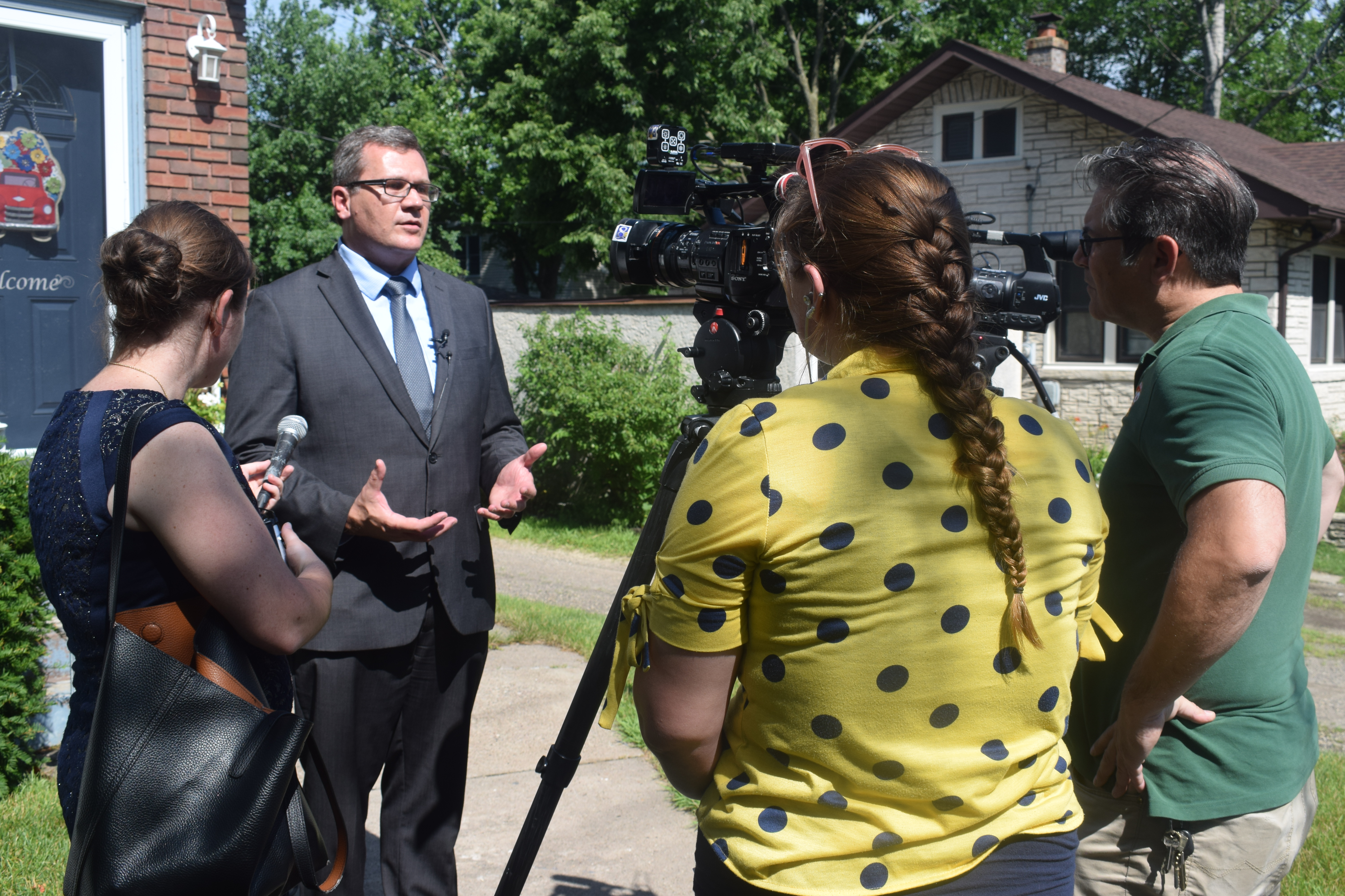 A man in a suit stands in front of a house surrounded by two reporters and a camera man. His hands gesture as he speaks with them.