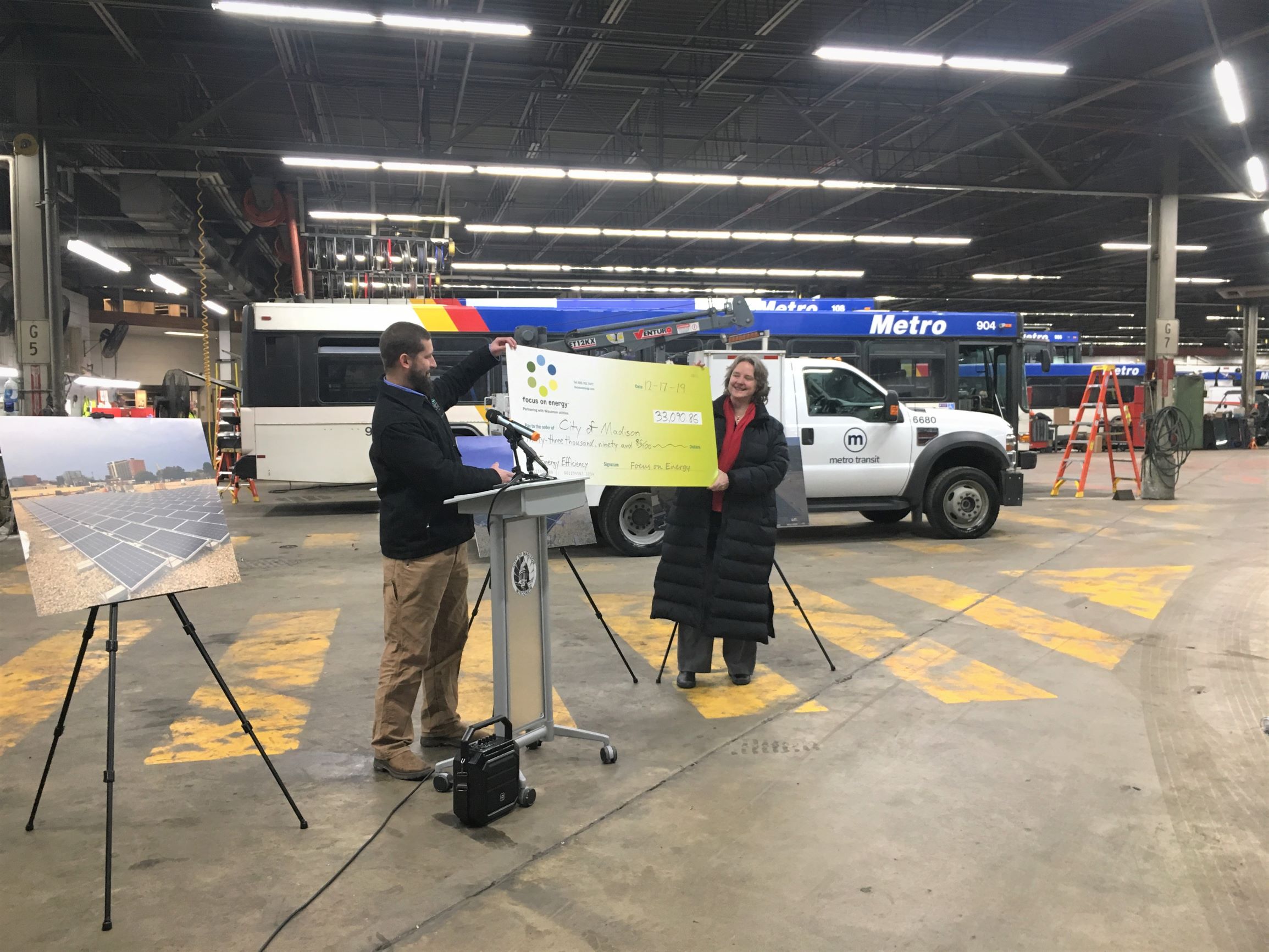 Two people stand holding a large, green ceremonial check with the Focus on Energy logo in a bus garage. A picture of solar panels is on display, and buses and other Metro Transit vehicles can be seen in the background. There is a podium in the center of the photo with the City of Madison seal. 