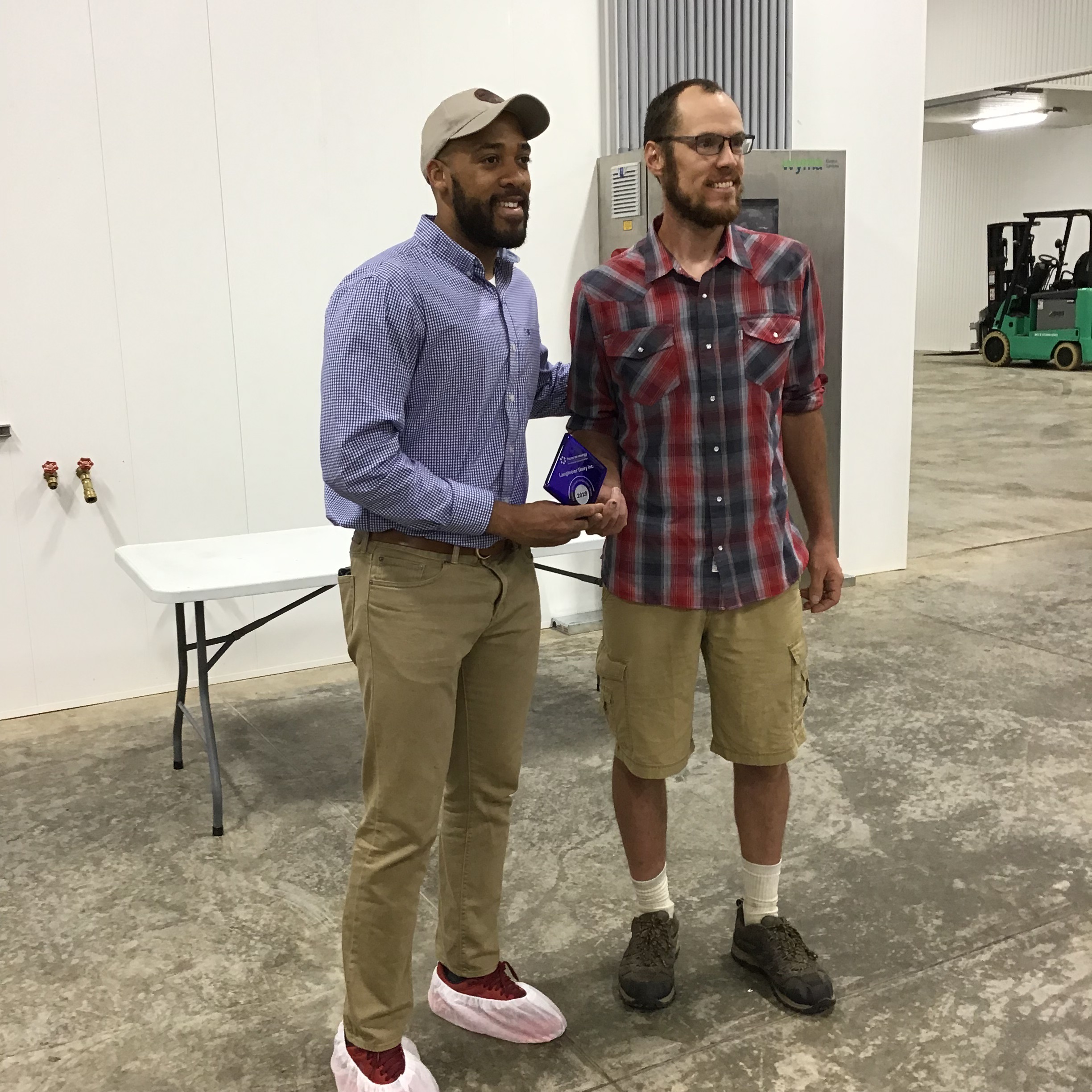 Two men stand in an industrial building, holding an award between them.