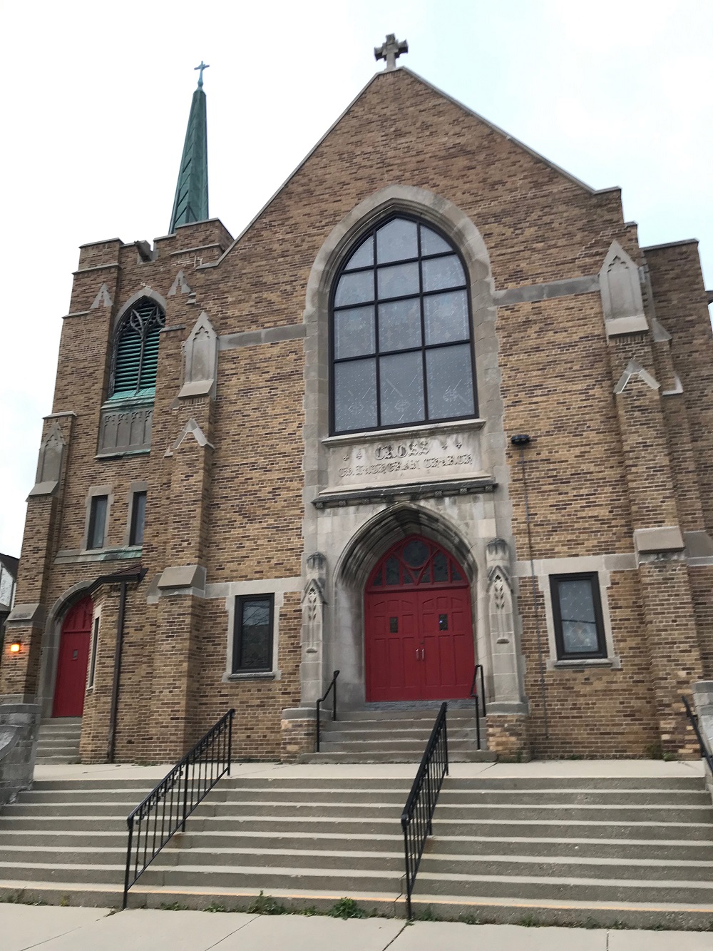 A brown brick church with red double doors and a green steeple and additional entrance with red doors to the left is pictured on an overcast day. There is a large stained glass window in the middle of the church, above the red doors.