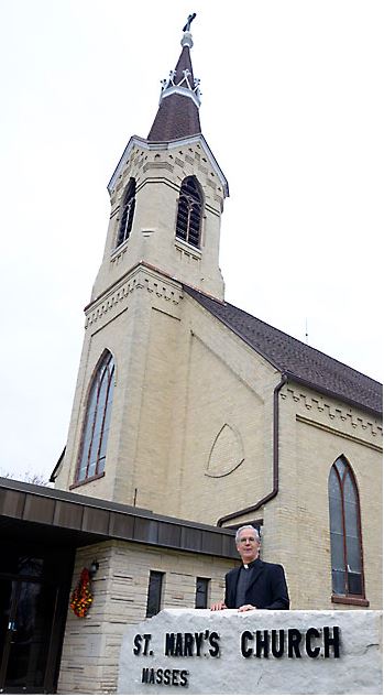 A priest stands in front of a beige brick building with a steeple. A stone sign reads "St. Mary's Church" with the word "Masses" under it. 